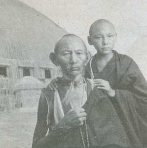 A black and white photograph depicting two Buddhist monks outside a straw building 