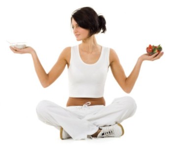 A woman sitting crossed legged holding a bowl of yogurt in one hand and a bowl of strawberries in the other